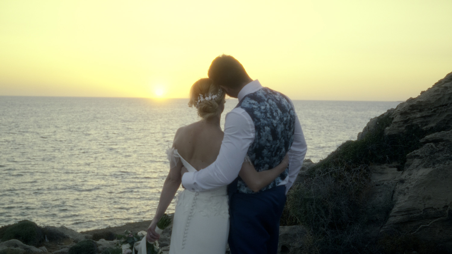Bride and groom standing on rocky coastline watching sunset over the Mediterranean Sea