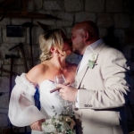Bride and groom kissing indoors at rustic wedding venue, holding wine glass, cigar, and bouquet
