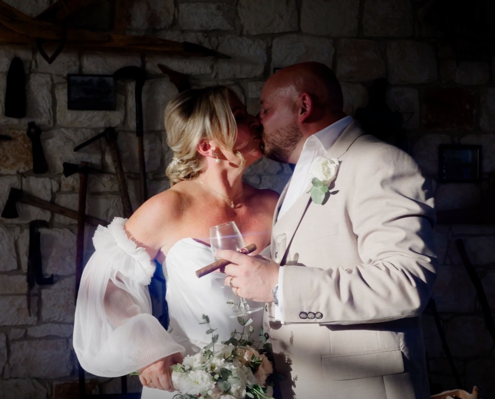 Bride and groom kissing indoors at rustic wedding venue, holding wine glass, cigar, and bouquet